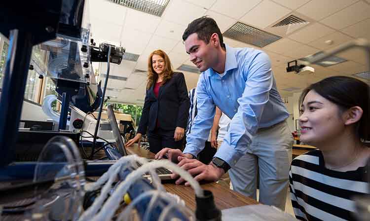 Mechanical engineering students are working in a small group with a professor smiling.