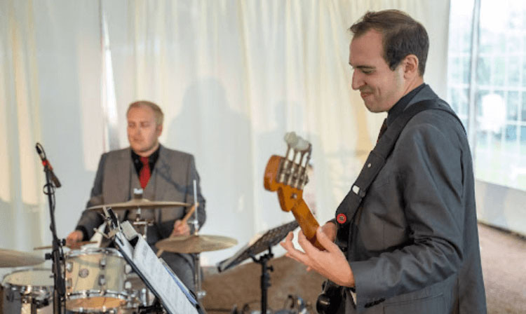 Two men playing music together wearing suits. The one in the back plays drums whereas the one in front reading sheet music plays bass guitar. 