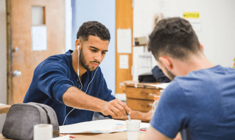 A student wearing wire headphones sits at a table and dips a paintbrush into a water cup.