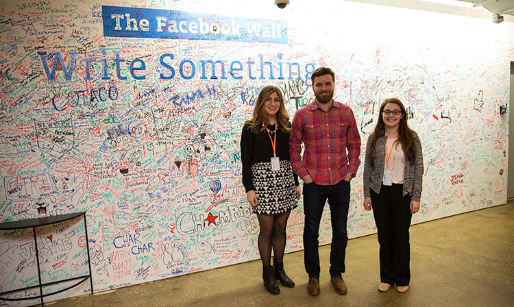 Three students standing in front of 'The Facebook Wall' during their Shadow a Stag experience.