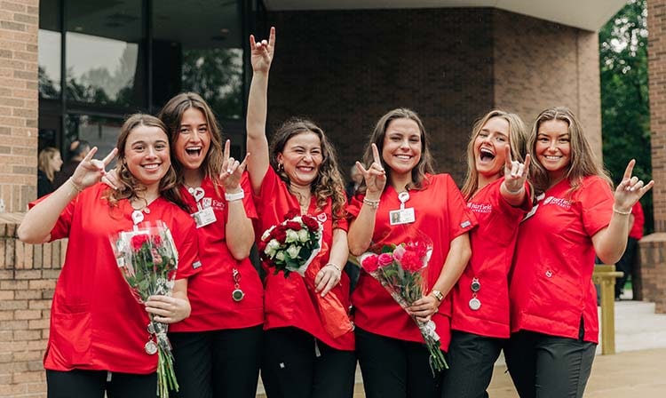 A group of students in red scrubs hold up their hands in a stag shape.