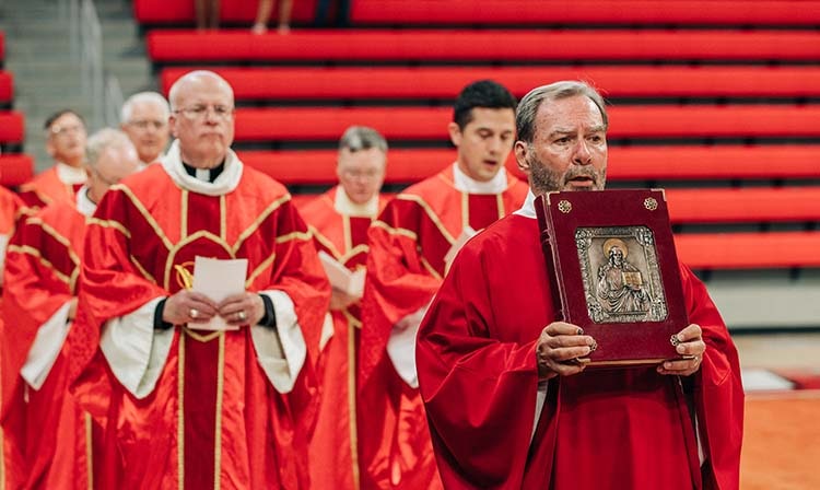 A priest holds a bible and stands in front of clergy.