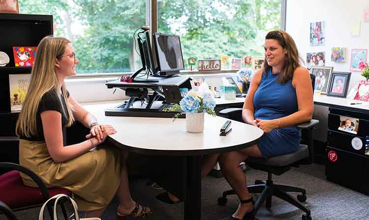 Image of two women chatting at a desk, looking at each other with friendly expressions.