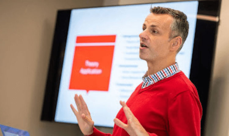 A professor in a red sweater stands in front of a PowerPoint addressing a class.