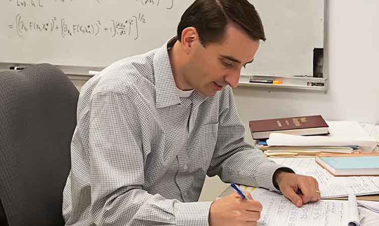 A professor sits at a desk looking at equations on a sheet of paper.
