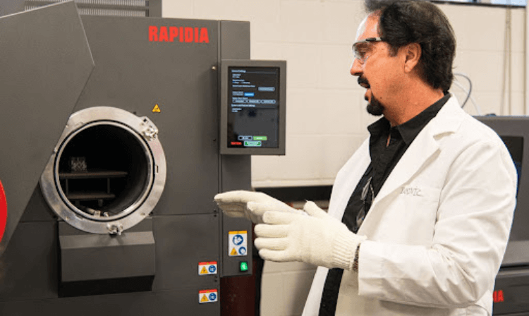 An engineering professor wearing a lab coat and eye protection explaining how a machine works in a lab. 
