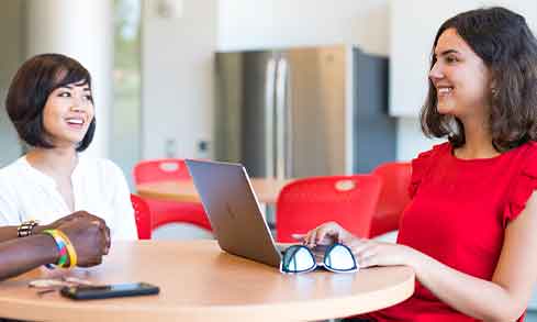 Two women engaged in conversation while sitting at a table with a laptop open between them.