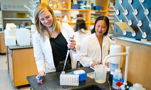 Two Biology students standing at a lab station. One of the students is using a dropper on a small test tube. 