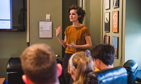 A professor addresses students in front of a PowerPoint screen.