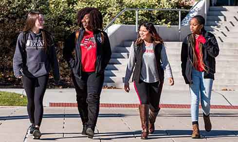 Four students walking on the sidewalk in front of a building, engaged in conversation and enjoying their time together.