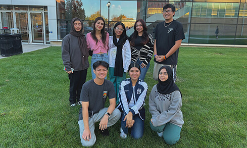 A diverse group of young students posing together on the grass outside a glass building.