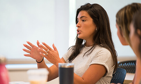 Two female students converse in a classroom, highlighting an interactive learning environment.
