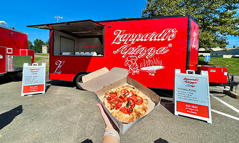 Student holding a personal margherita pizza in a box, made fresh and to order by the Zuppardi's Apizza food truck.