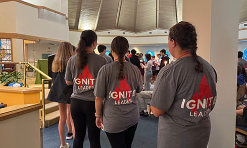 Three staff members in gray GNIT Leader shirts stand in a bright event hall, facing a busy crowd; the scene feels professional and collaborative.