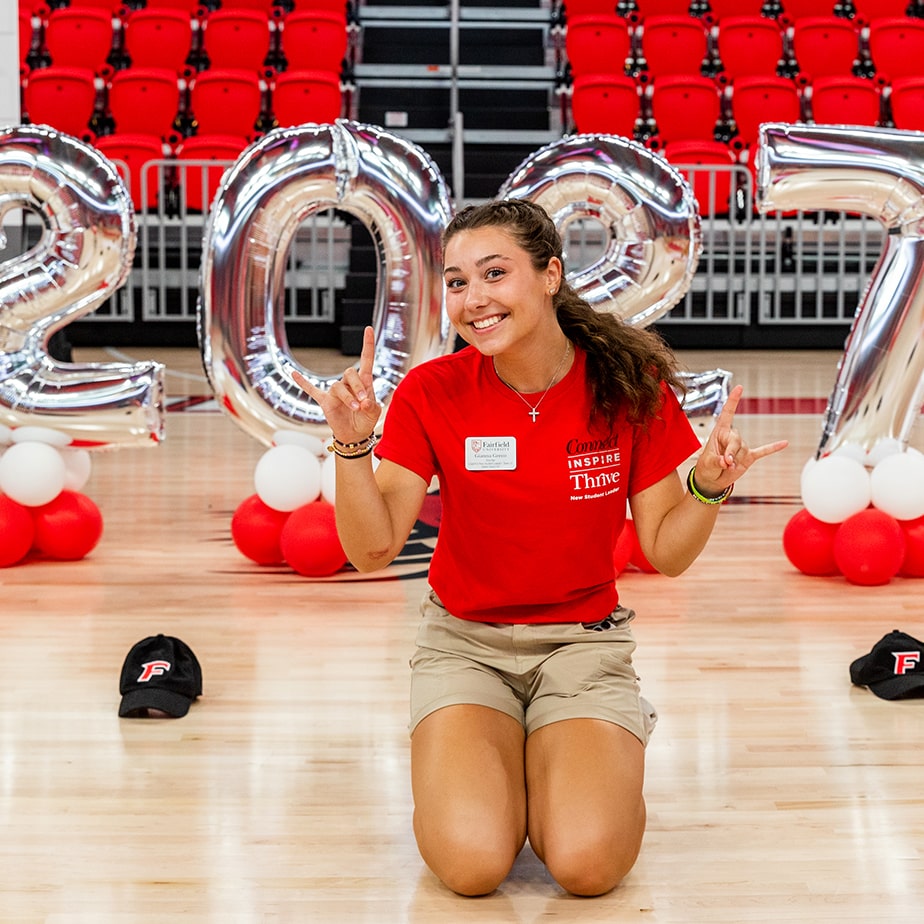 A student kneels on the ground in front of silver balloons that read 2027.