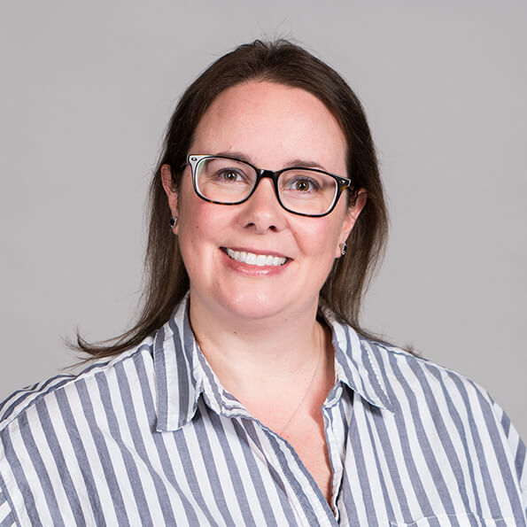 A woman with glasses and long brown hair smiles warmly. She wears a striped shirt against a neutral gray background, conveying a friendly, professional tone.