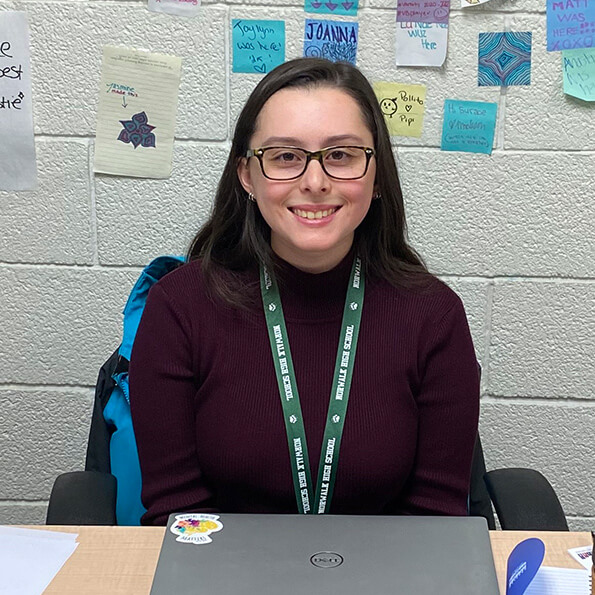 Emily Sgritta '26 with glasses and a green lanyard sits behind a laptop, with a blue backpack and colorful handwritten notes on the wall behind her.