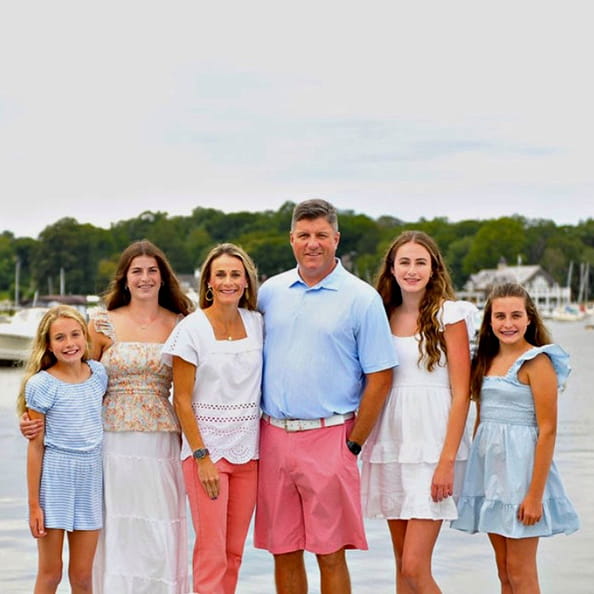 A family of six, dressed in light summer clothing, poses happily by a calm lake with boats. Lush greenery in the background under an overcast sky.