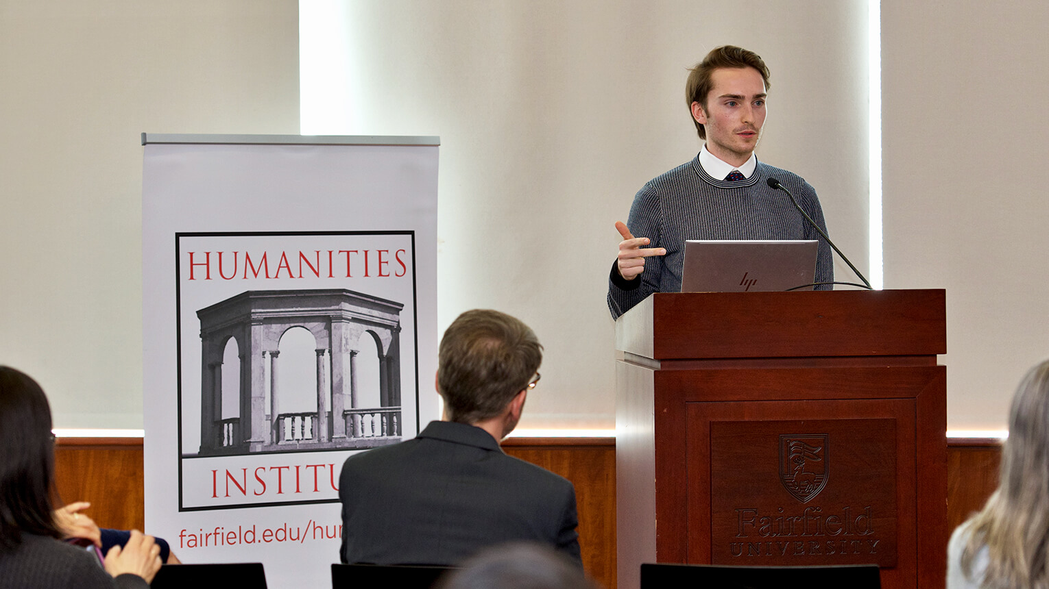 A student speaker presents at a podium while a crowd attentively listens in a well-lit auditorium.
