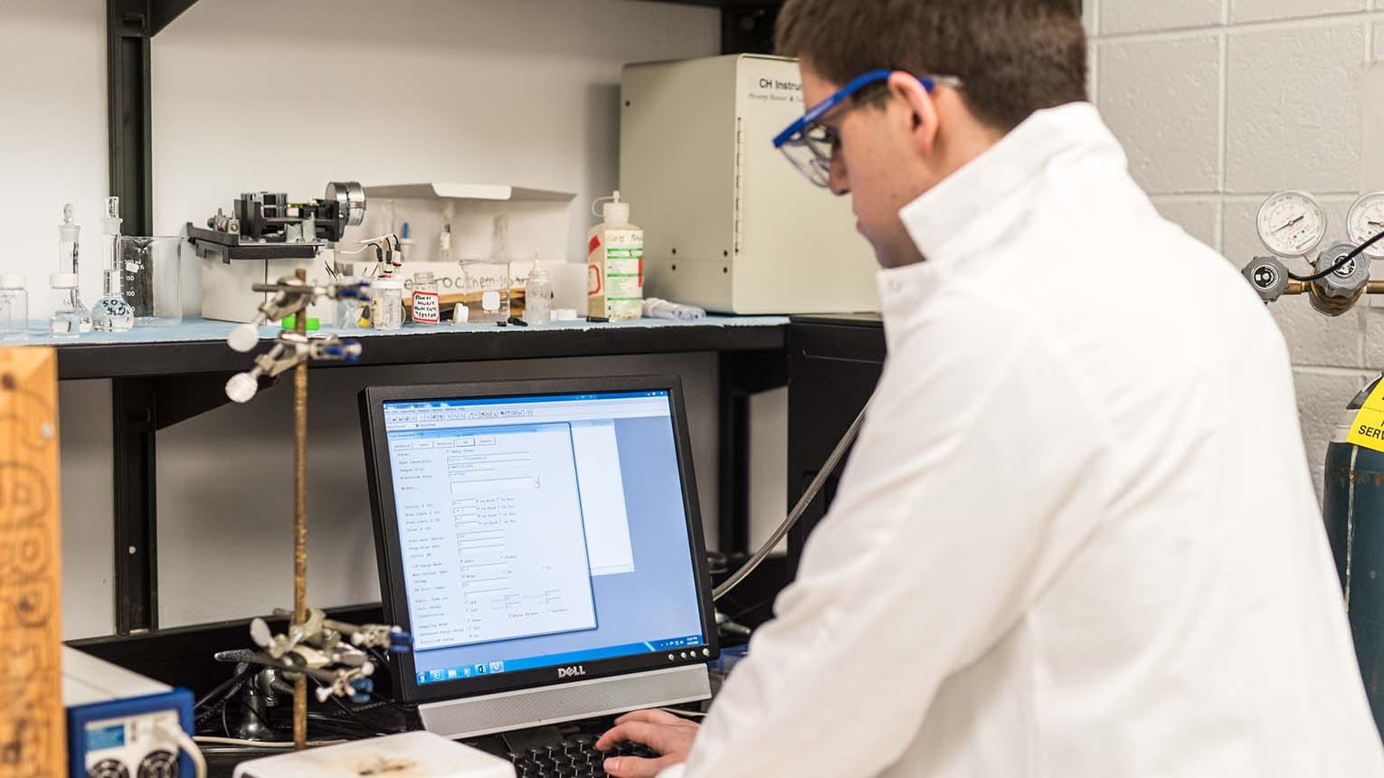 Engineering student in protective eyewear and a lab coat performing an experiment where he measures cyclic voltammetry.