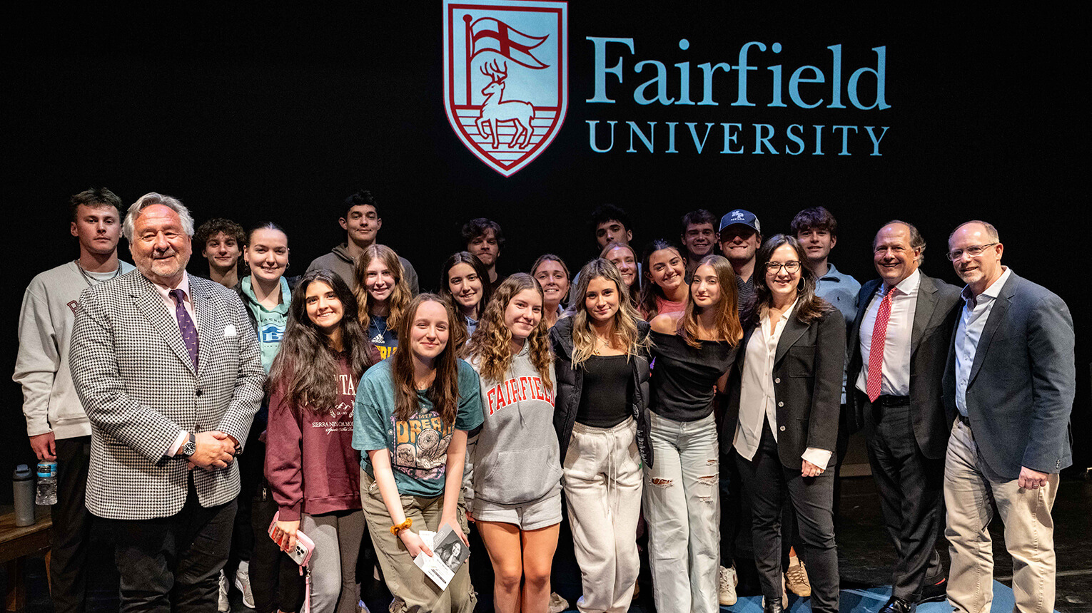 A group of students, faculty and Bari Weiss pose on stage with smiles, the Fairfield University logo displayed behind them. The atmosphere is cheerful and collegiate.