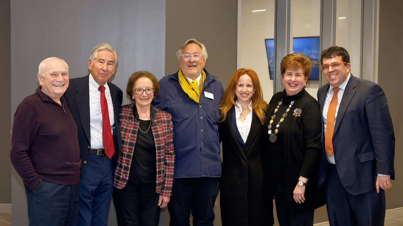 A group of seven people stand closely together, smiling warmly. They wear formal and semi-formal attire, exuding a friendly and celebratory tone.