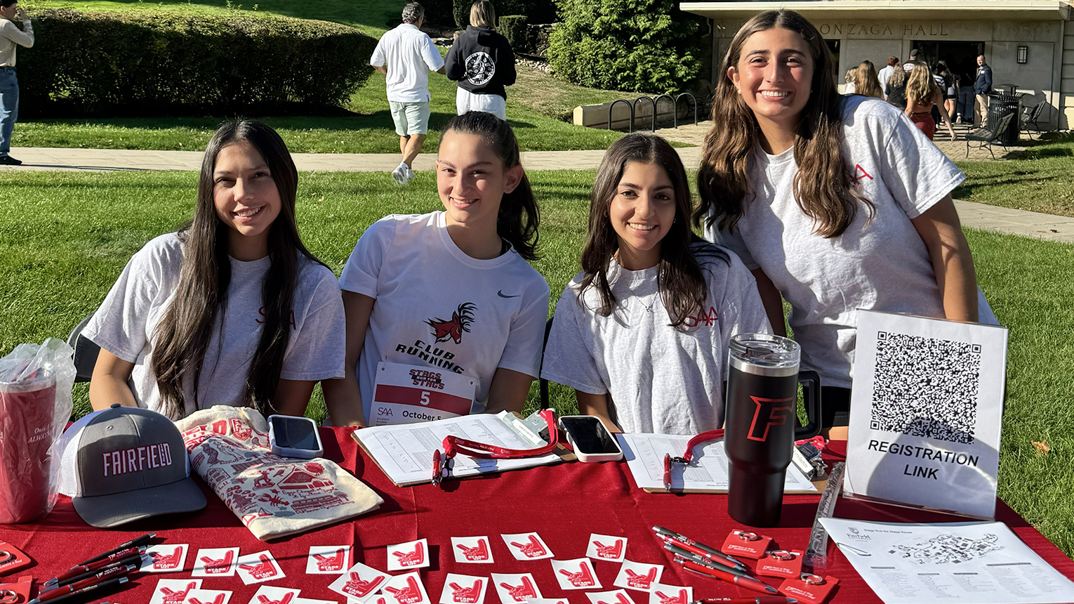 Four smiling young women sit at a red outdoor registration table, surrounded by promotional items and greenery in the background.