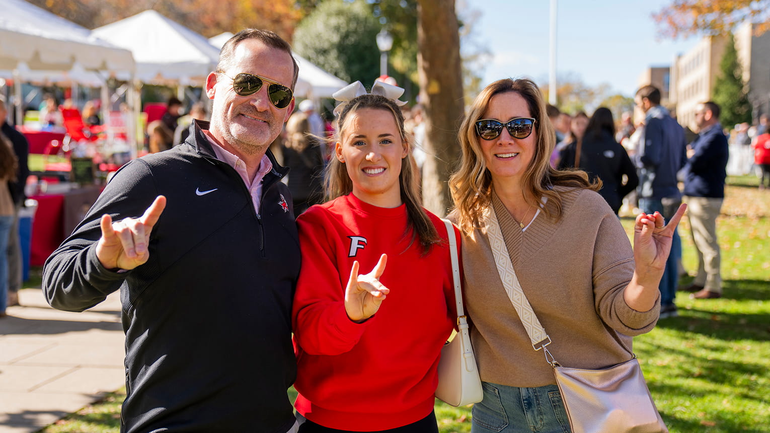 A group of three smiling adults stands outdoors on a sunny day, making hand gestures. They're dressed casually, with a crowd and tents in the background.