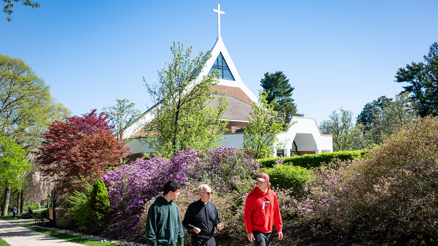 Three people walk beside a modern white church with a cross, surrounded by blooming spring shrubs and trees under a clear blue sky.