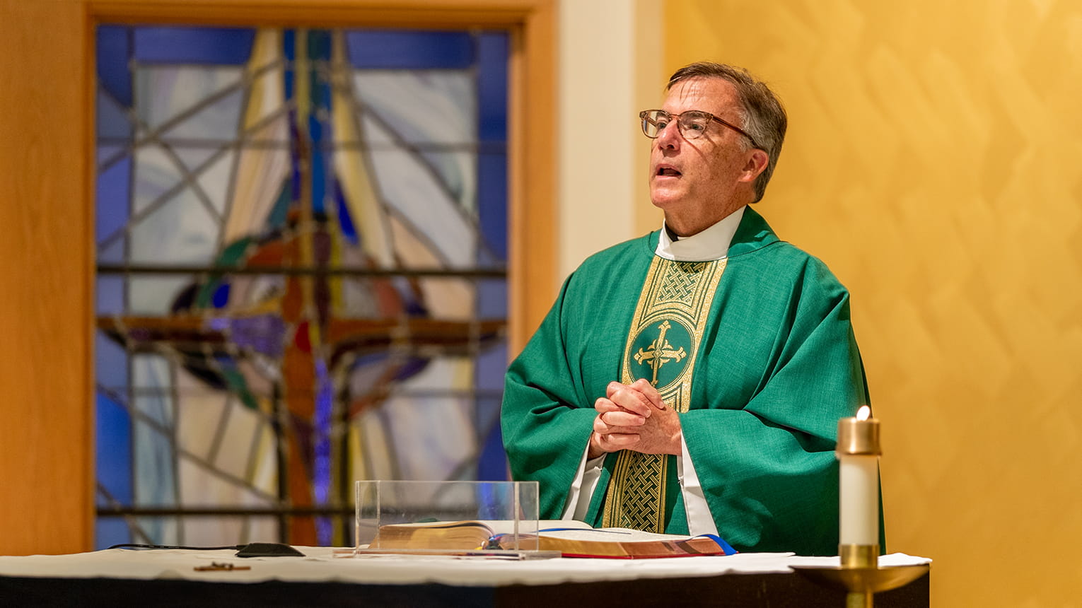 Priest in green vestments stands at an altar with clasped hands, speaking reverently before a colorful stained-glass cross.