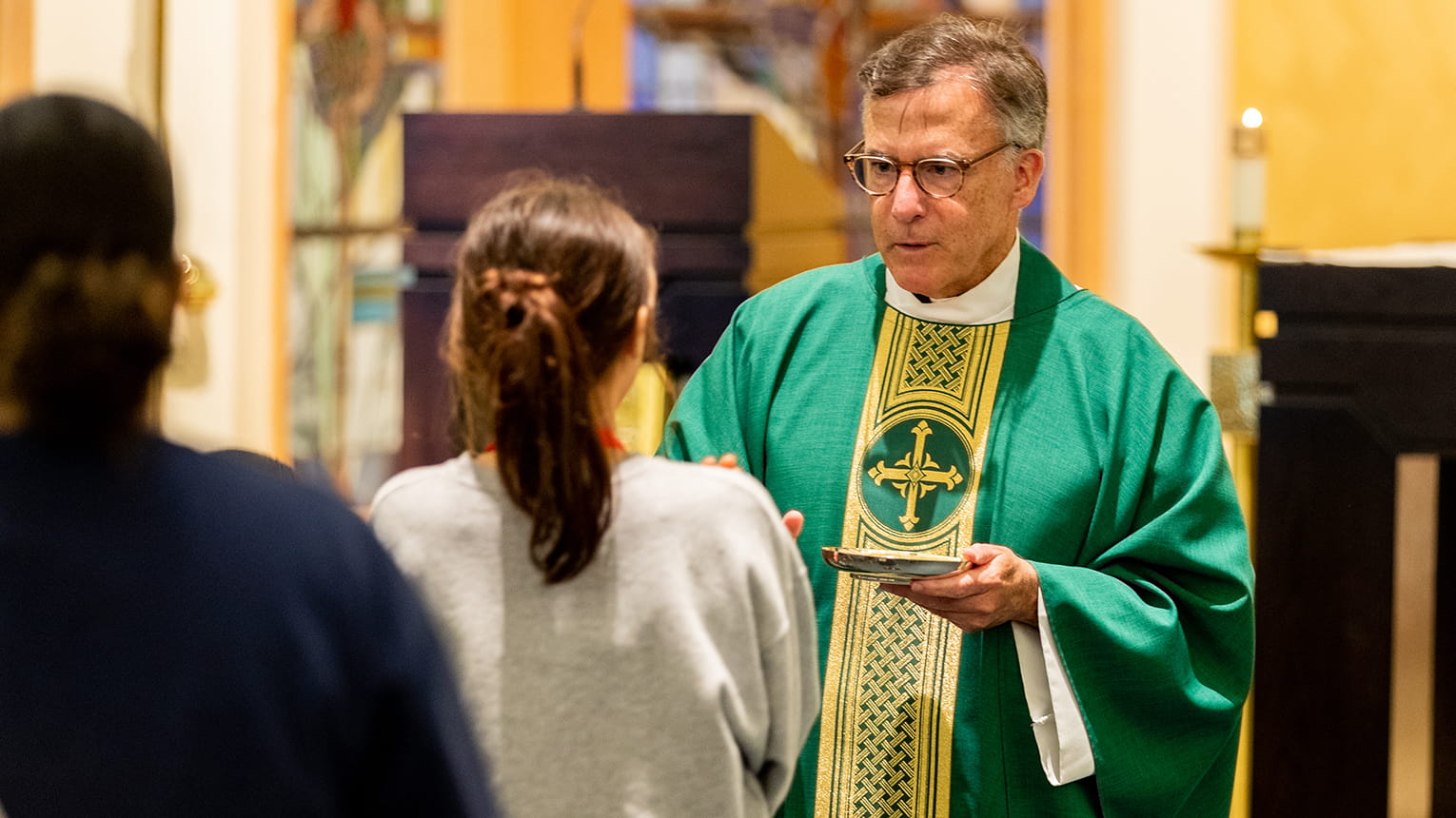 Catholic priest in green vestments holds a communion plate before a woman in church, with another congregant nearby; solemn, warm scene.