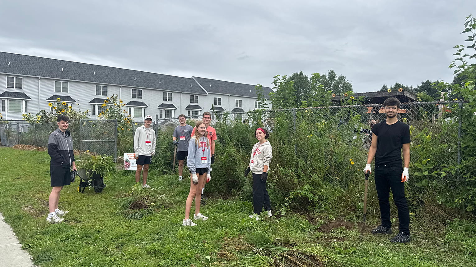 Five students and an adult stand beside a fenced school garden, with raised beds, greenery, and a hoop visible; the overcast scene feels calm and educational.