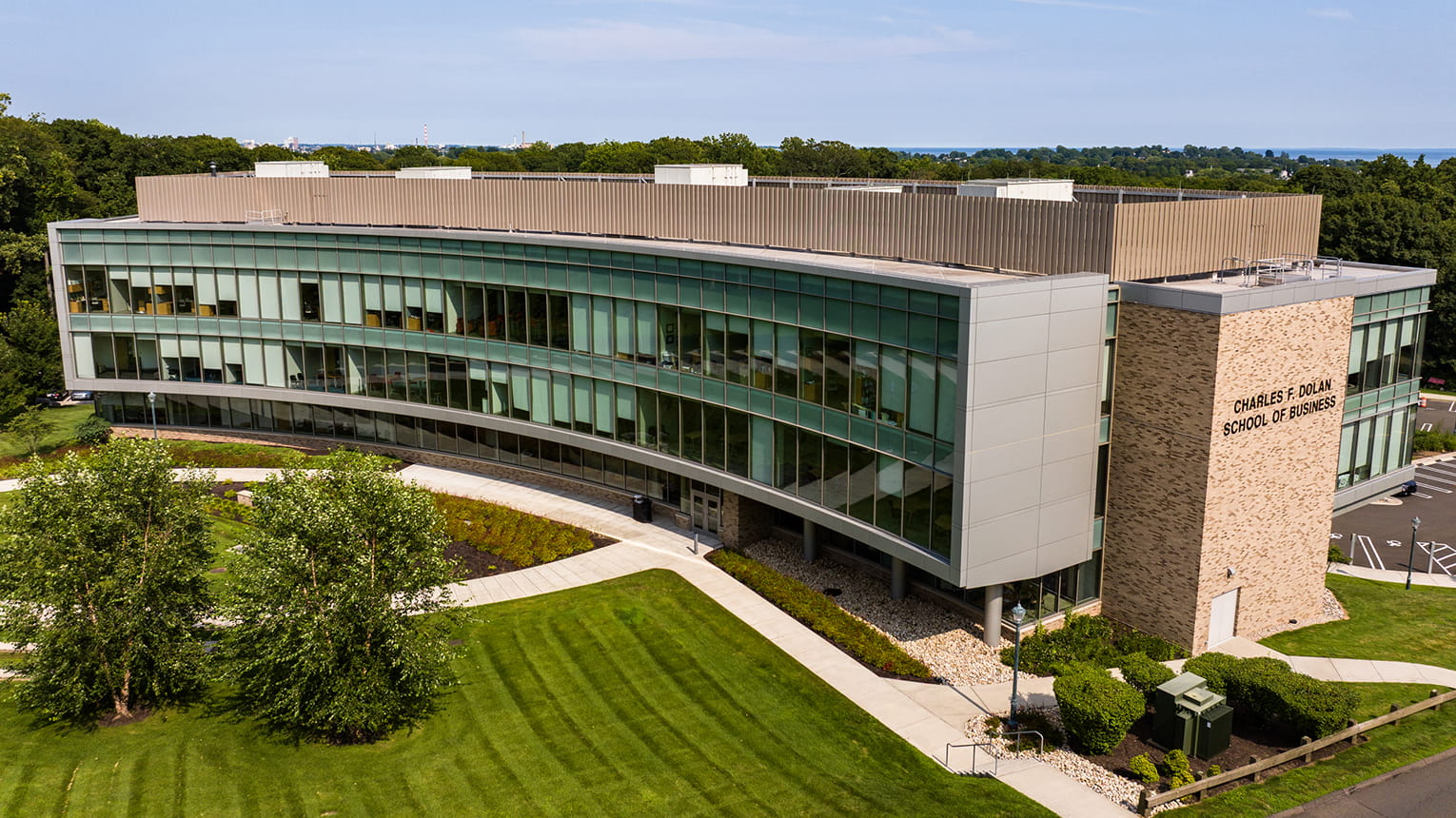 Aerial view of a modern curved glass campus building with a stone entrance tower, surrounded by green lawns, trees, and walkways; calm and welcoming.