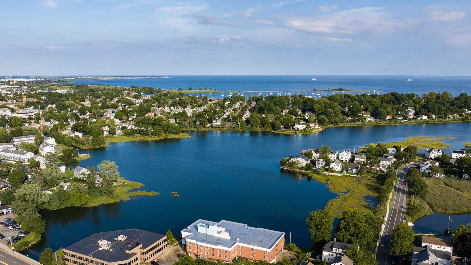 Aerial view of tranquil blue water surrounded by leafy neighborhoods, homes, and a road, with the open sea and pale sky beyond.
