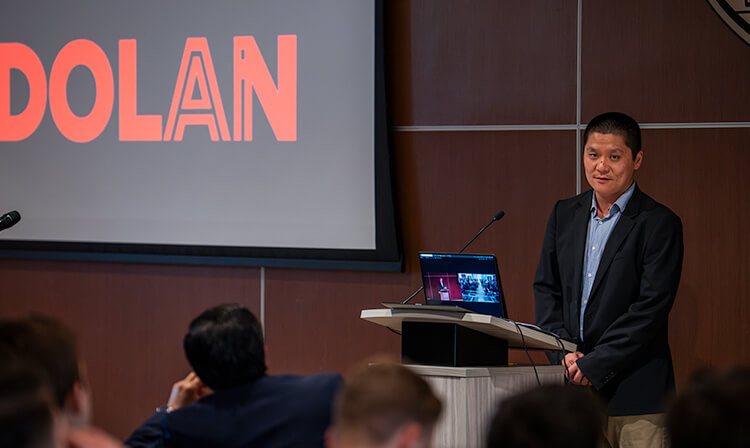 A man in business attire stands before a podium, with a laptop open in front of him.