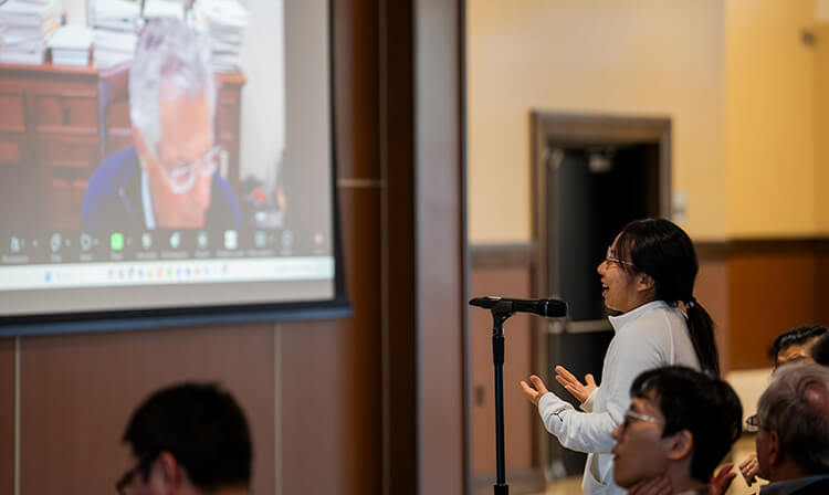 A woman speaks into a microphone, addressing an audience in front of a large presentation screen.