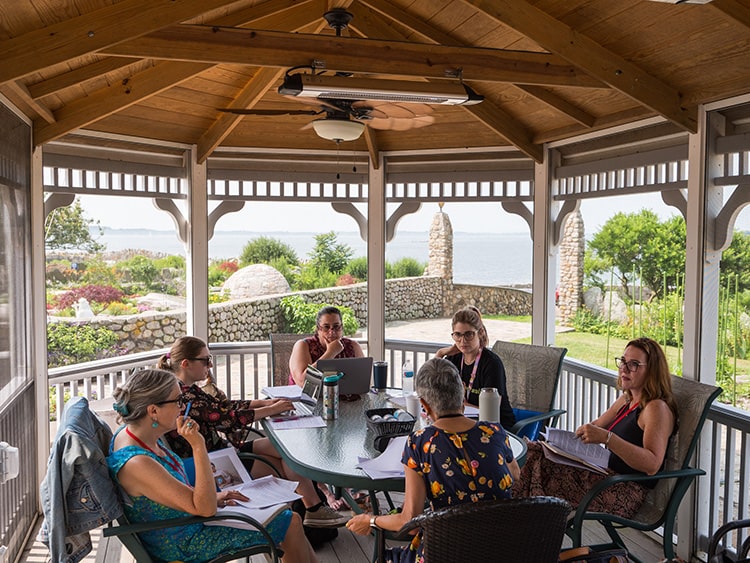 Creative writing MFA candidates collaborating in an intimate group setting under a gazebo on a bright summer day.