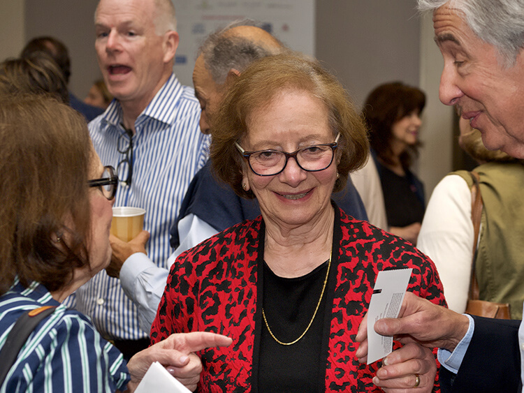 Debby Zieff in a red and black jacket smiles warmly during a social gathering. She engages with two people holding a paper. The mood is lively and cheerful.