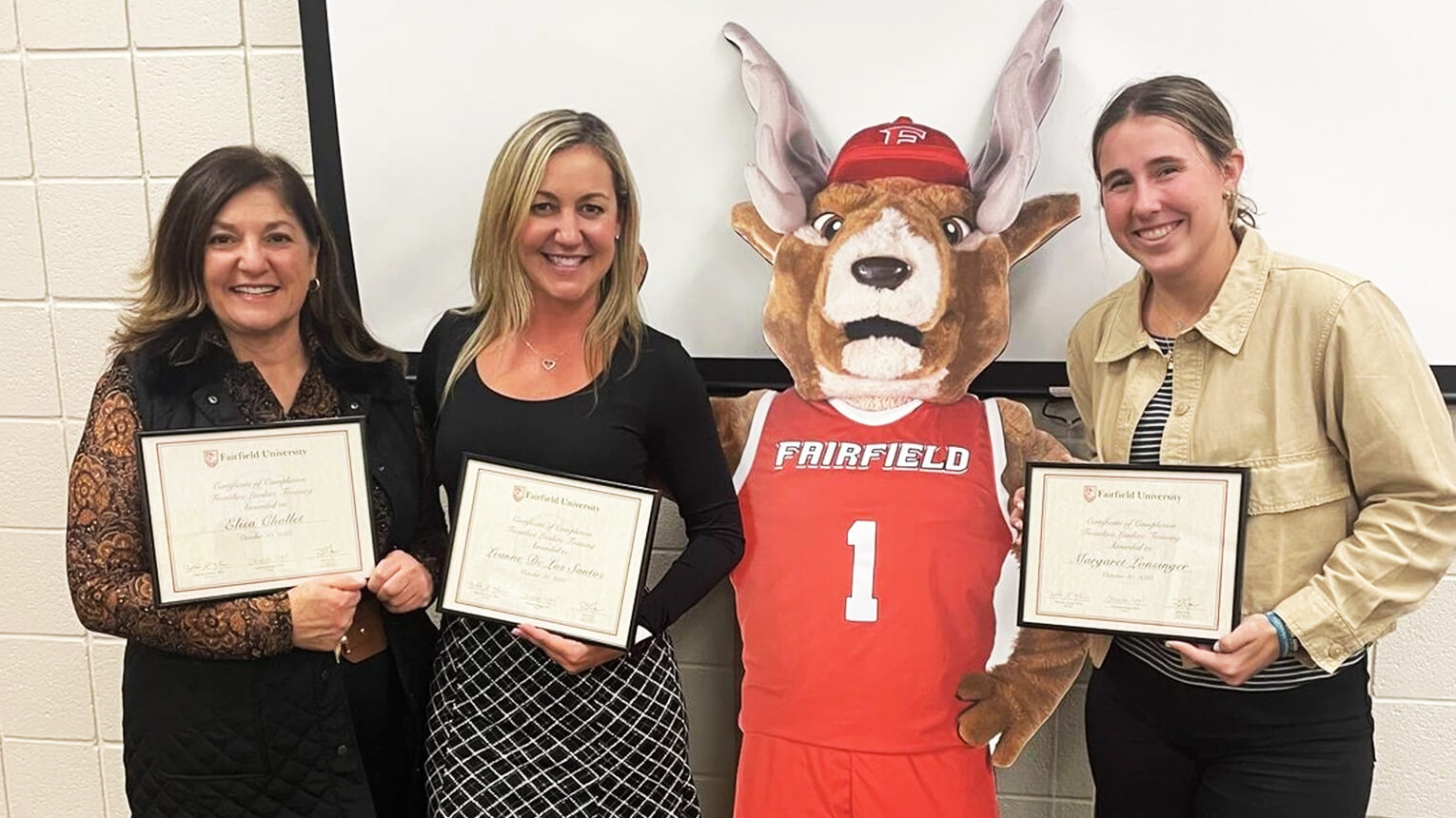 Three people smile while holding certificates beside Lucas the Stag mascot in a red jersey. The setting is a classroom with a positive, celebratory tone.