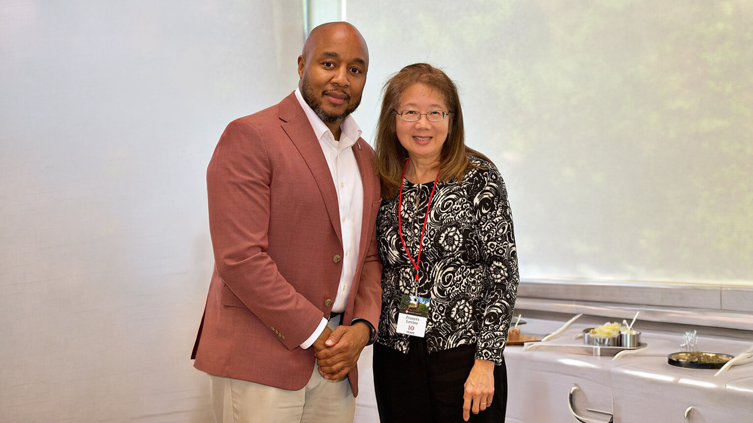 A man in a maroon blazer and a woman in a patterned blouse smile at the camera in a bright room. They stand by a buffet table, creating a warm, welcoming atmosphere.