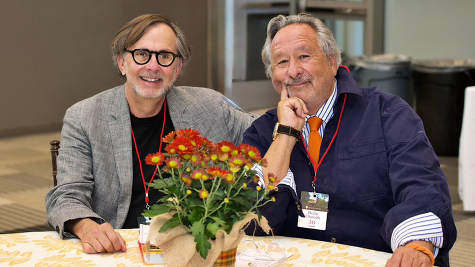 Two men seated at a table with a vibrant potted plant centerpiece, both smiling and wearing lanyards. The setting appears casual and friendly.