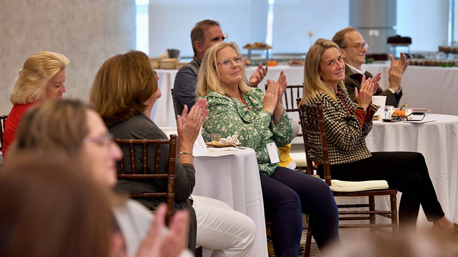 A group of people at a seminar clapping, seated around tables. They appear attentive and pleased, creating a positive and engaged atmosphere.