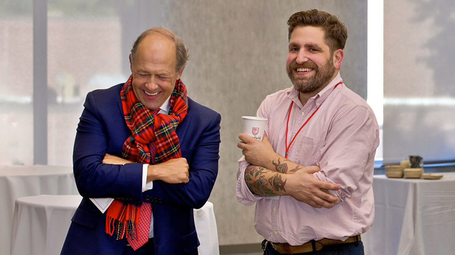 President Mark Nemec and another man stand in a bright room, both smiling warmly. One wears a blue suit and red plaid scarf, the other a pink shirt with tattoos, holding a coffee cup.