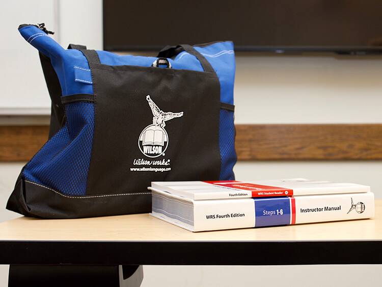 A Wilson-branded tote bag with blue accents sits on a desk beside two thick manuals titled "WRS Fourth Edition" and "Instructor Manual." The setting is educational.