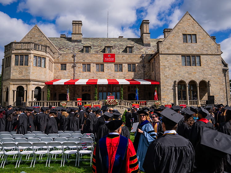 Graduates in black caps and gowns gather before a historic stone building, Bellarmine Hall, with red awnings, flower displays, and rows of empty chairs.