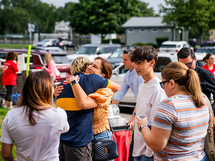 People, including a mother and son, warmly embrace in a busy outdoor parking lot, surrounded by families, cars, and trees—a joyful, welcoming community moment.