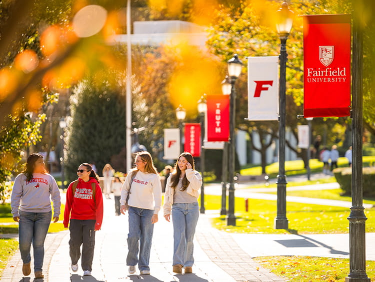 Four Fairfield University students walk along a tree-lined campus pathway during a sunny autumn day with Fairfield banners displayed on lamp posts.