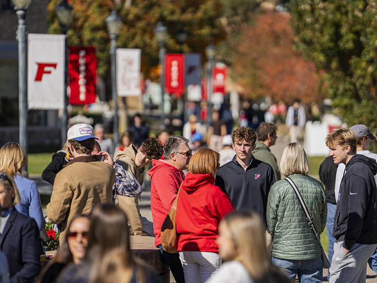 Families and students gather along a campus walkway during Fairfield University Family Weekend with fall foliage and campus banners in the background.