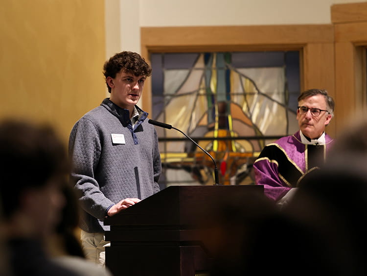 Young man speaks at a church podium beside a priest in purple vestments; a stained-glass window and blurred audience create a solemn tone.