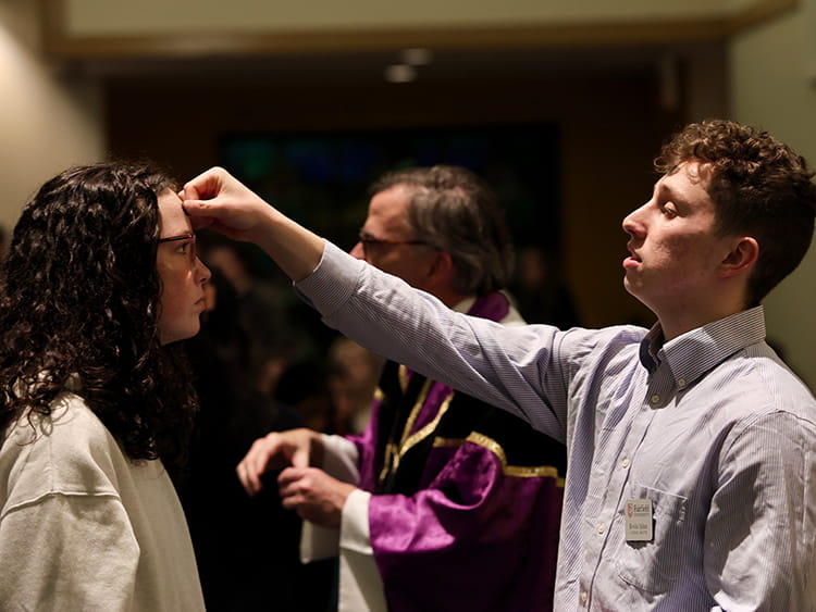 A young man places ashes on a woman’s head during a liturgical ceremony, while a robed priest stands behind them; warm, attentive atmosphere.